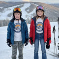 Two children in ski gear standing on a snowy slope with mountains in the background.